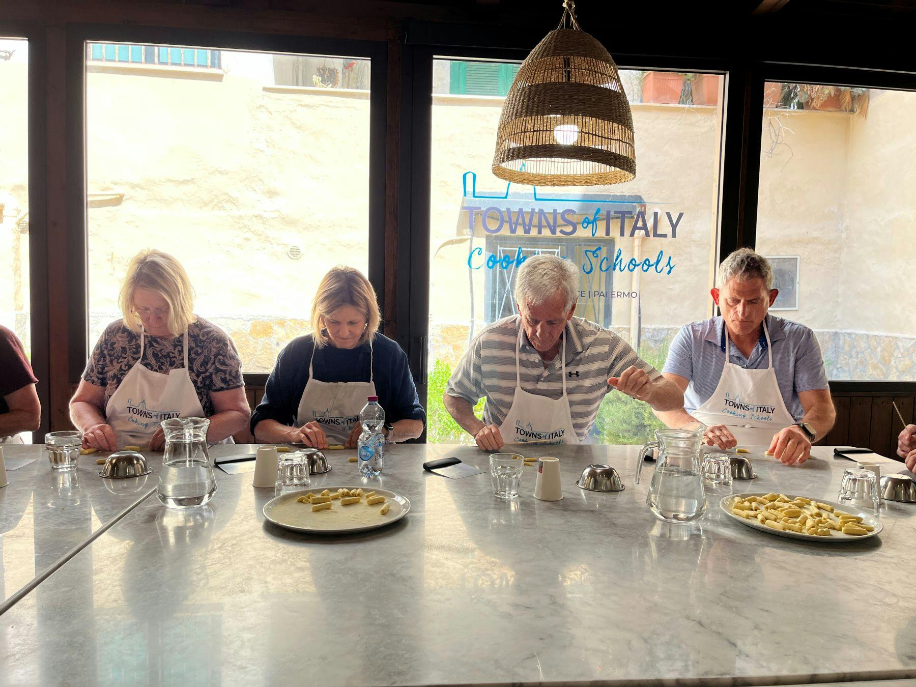 Three people seated at a cooking class table with aprons, preparing food with utensils, near a plate of pasta.