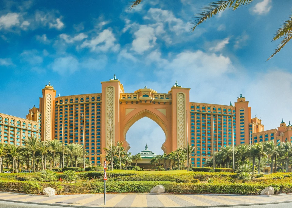 A large, luxury hotel with an archway in the center, surrounded by palm trees and lush landscaping, under a partly cloudy sky.