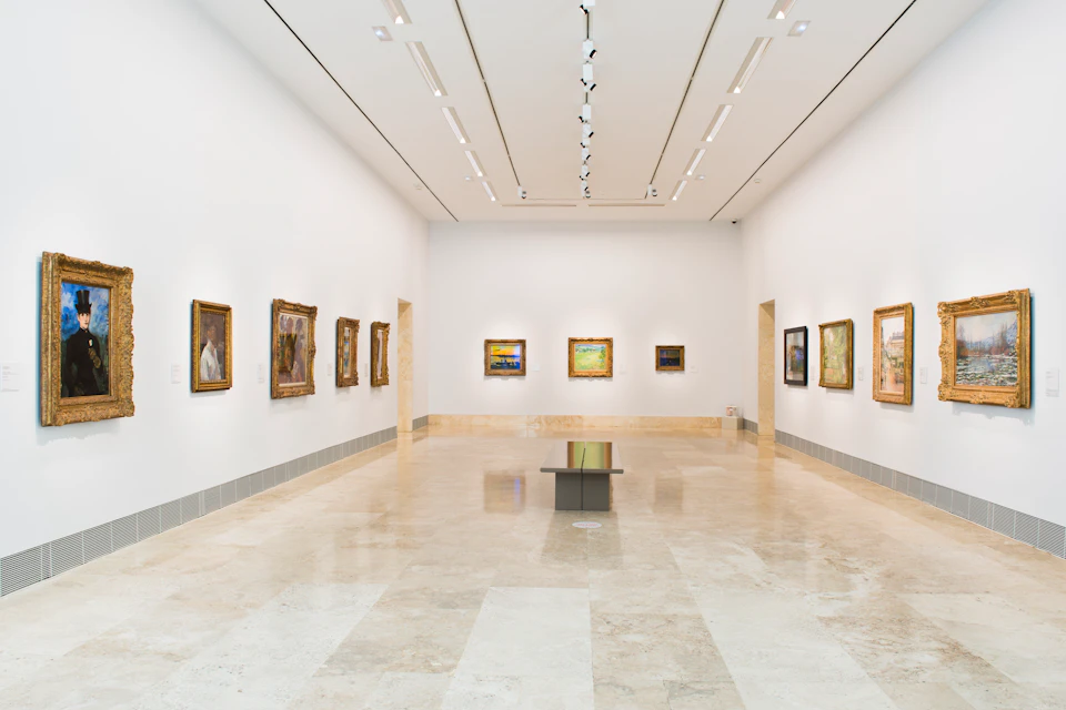 An empty art gallery with framed paintings on white walls, a marble floor, and a central bench. Wall-mounted spotlights illuminate the artwork.