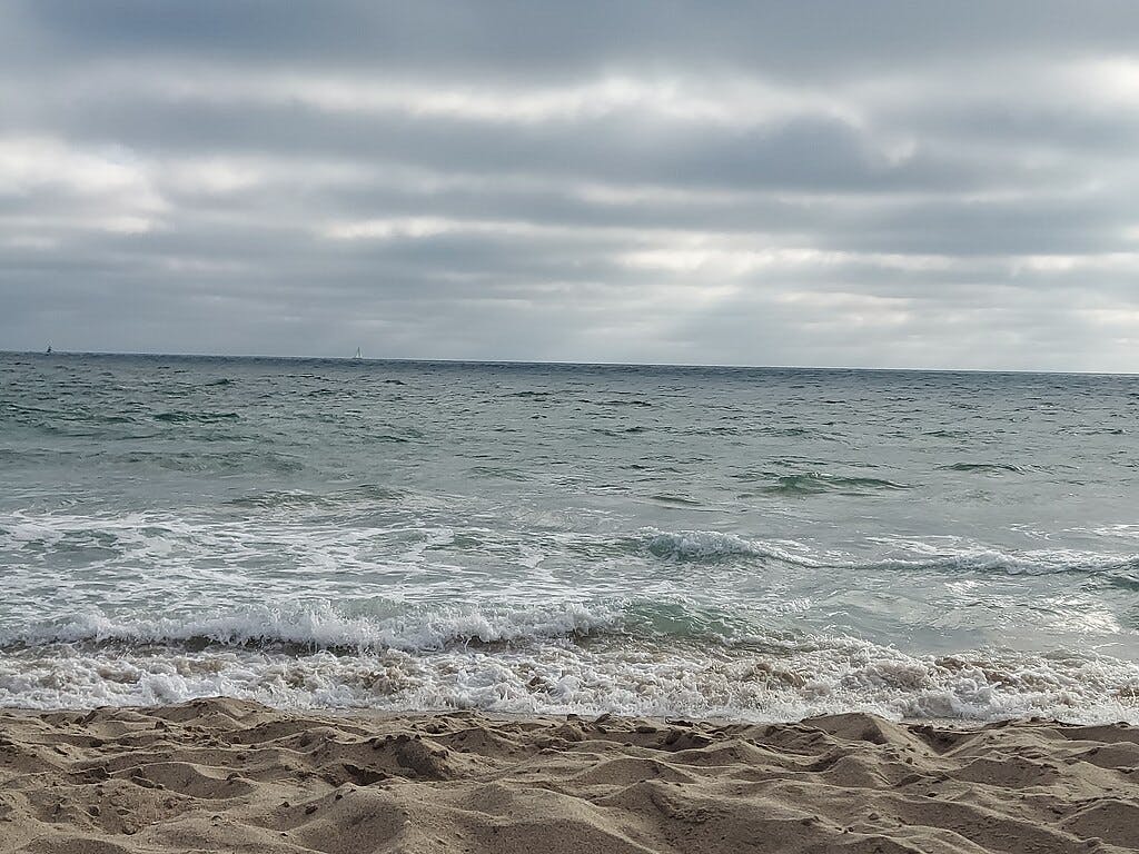 Cielo nublado sobre un océano en calma con suaves olas que bañan una playa de arena.