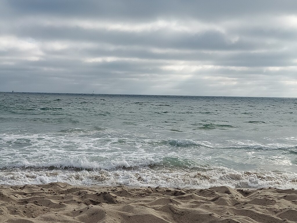 Cel ennuvolat sobre un oceà tranquil amb suaus onades que s'estenen sobre una platja de sorra.
