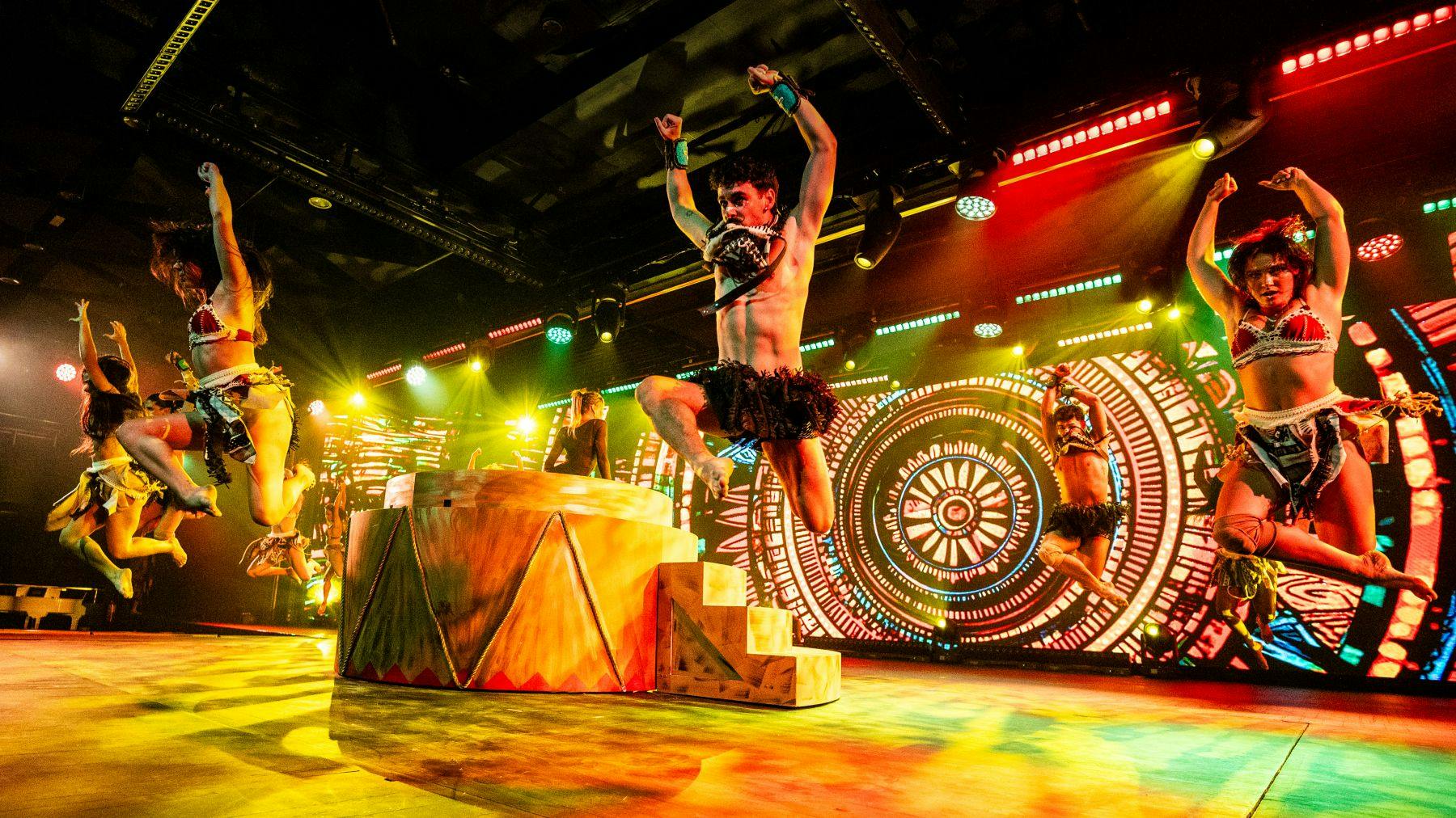 Um espetáculo dinâmico em palco com bailarinos em trajes de inspiração tribal e iluminação colorida contra um pano de fundo circular.