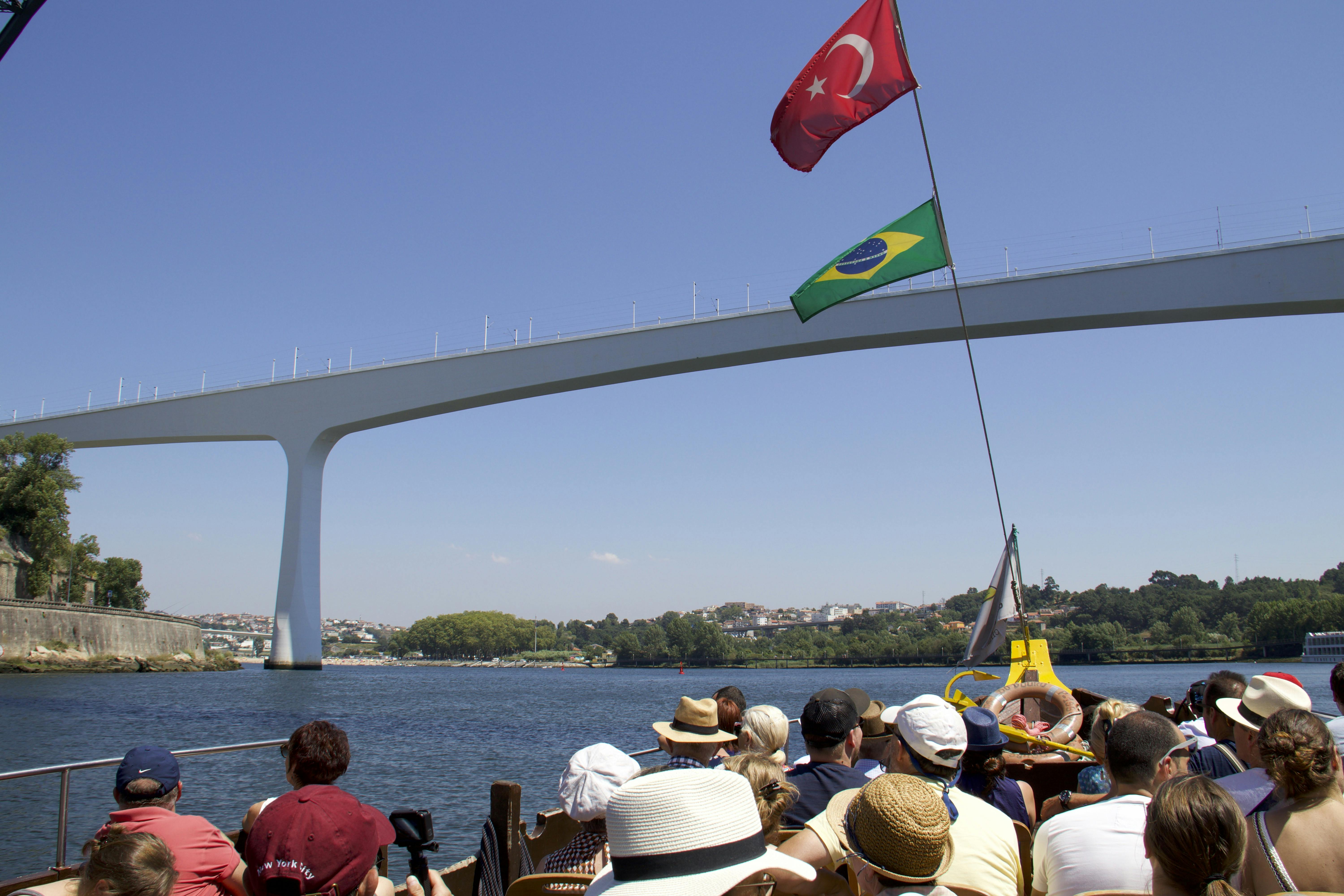 Ludzie na wycieczce łodzią pod nowoczesnym mostem łukowym z tureckimi i brazylijskimi flagami na tle czystego błękitnego nieba.
