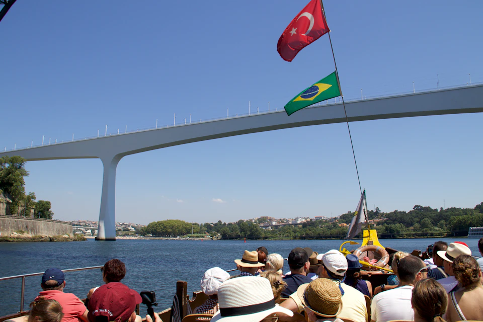 People on a boat tour under a modern arch bridge with Turkish and Brazilian flags against a clear blue sky.