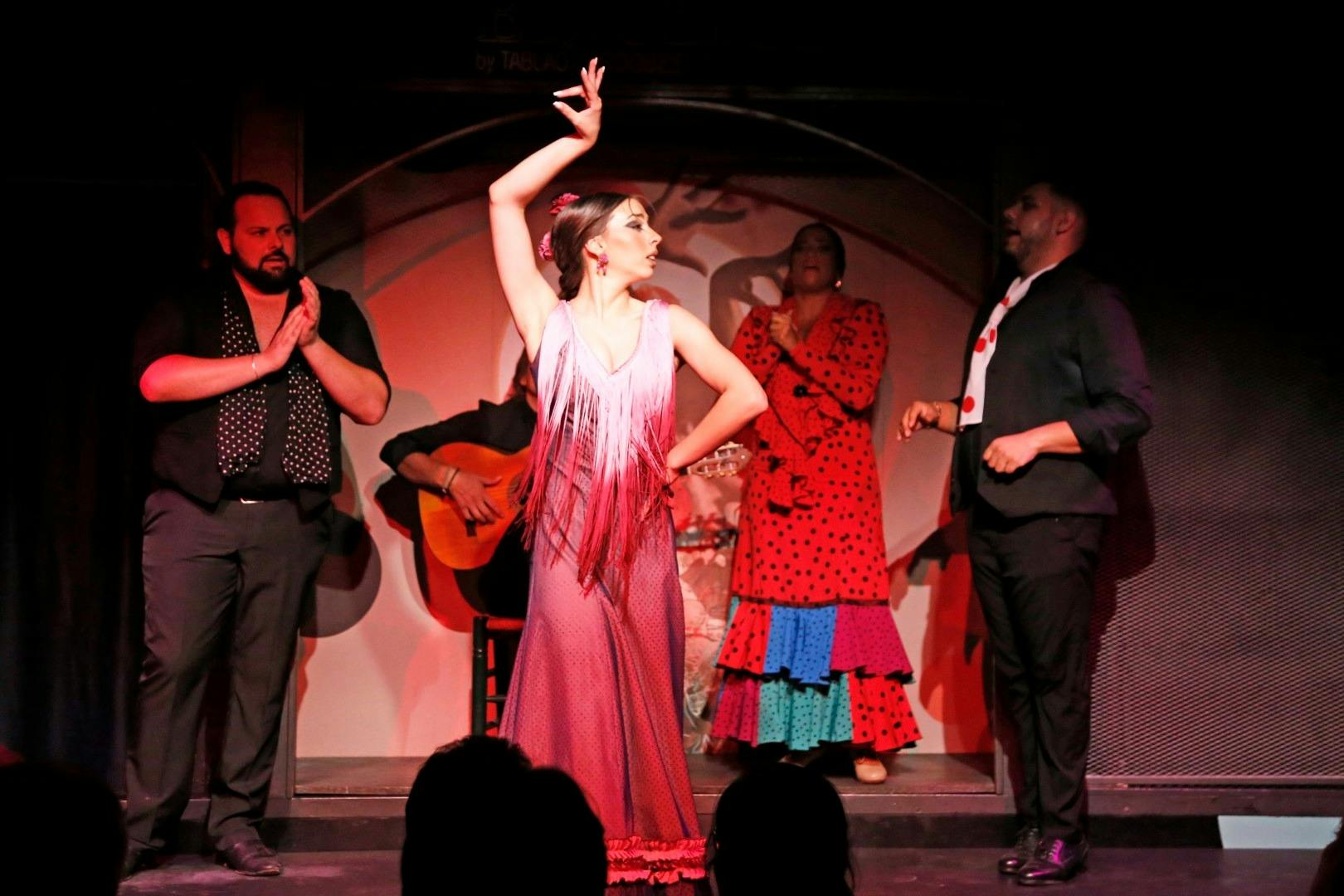 Een flamencovoorstelling met een danseres in een roze jurk, begeleid door een gitarist en kleppers op een podium.