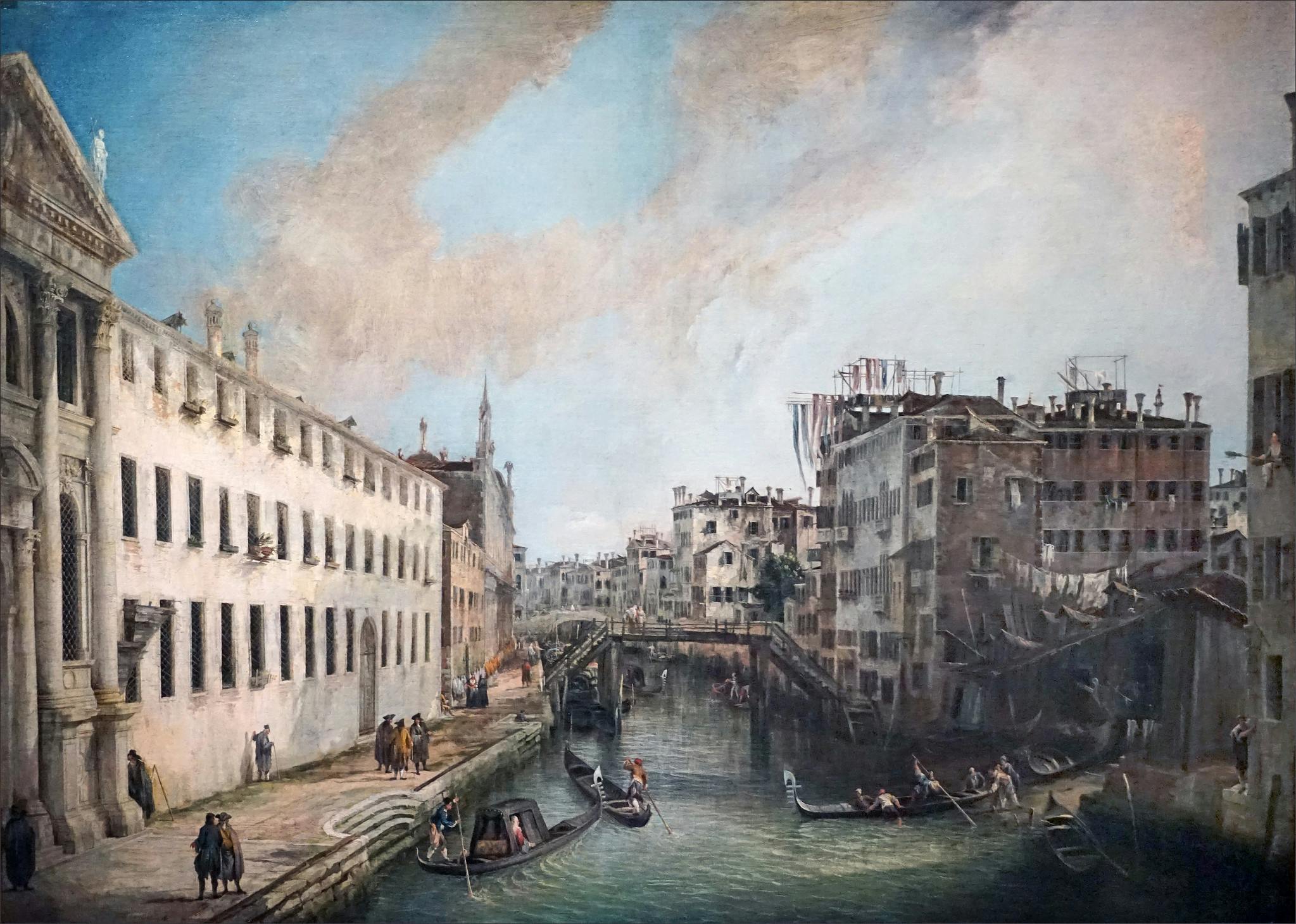 A historic cityscape with gondolas on a canal, buildings on both sides, and people walking along the waterfront.