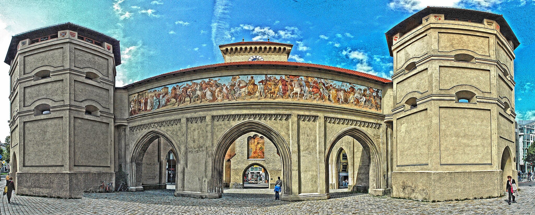 Historická kamenná brána s oblouky, barevná nástěnná malba zobrazující historické postavy, dvě osmiboké věže a procházky lidí.