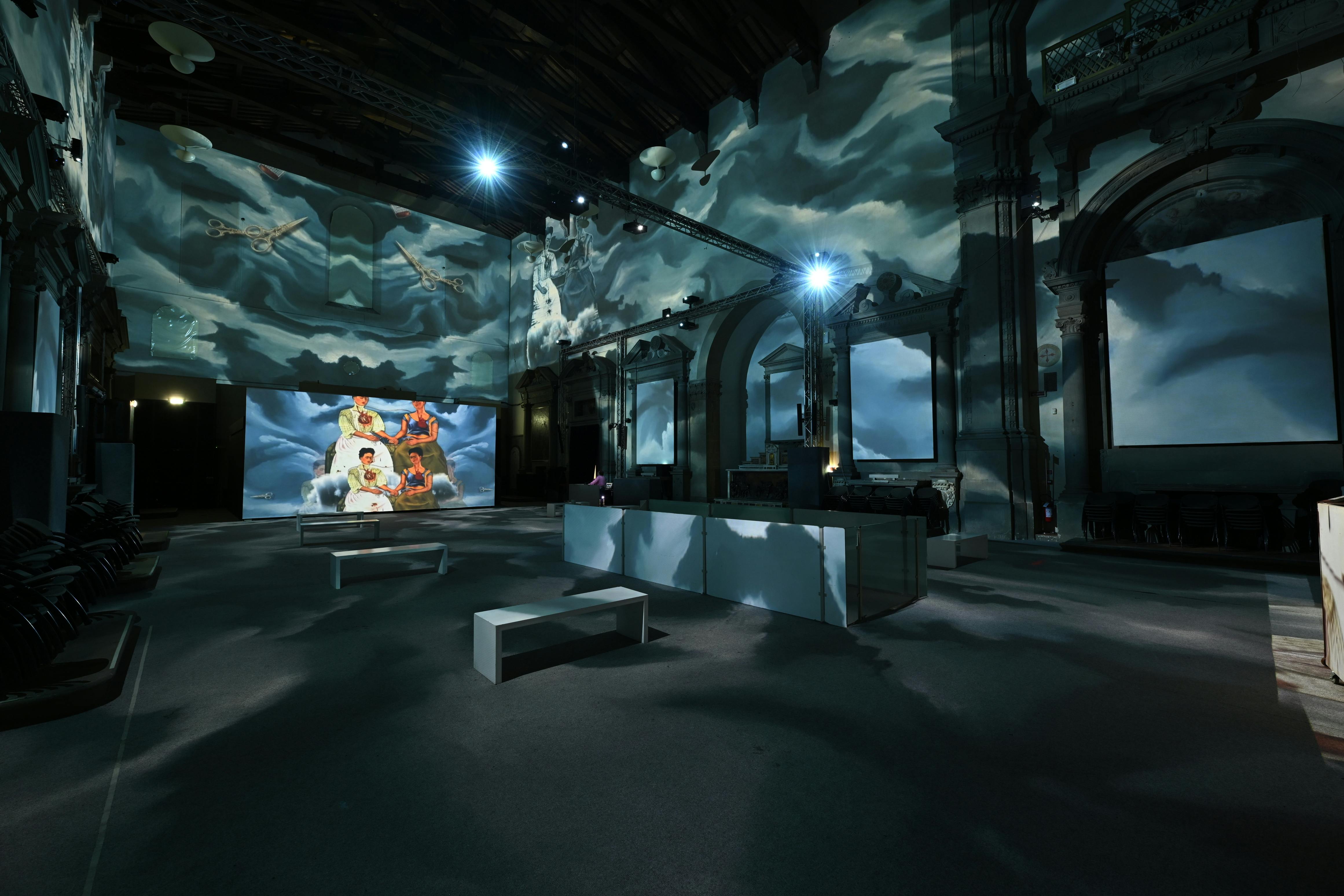 A dimly lit room with projected cloud imagery on walls and ceiling, featuring benches and a screen displaying artwork.