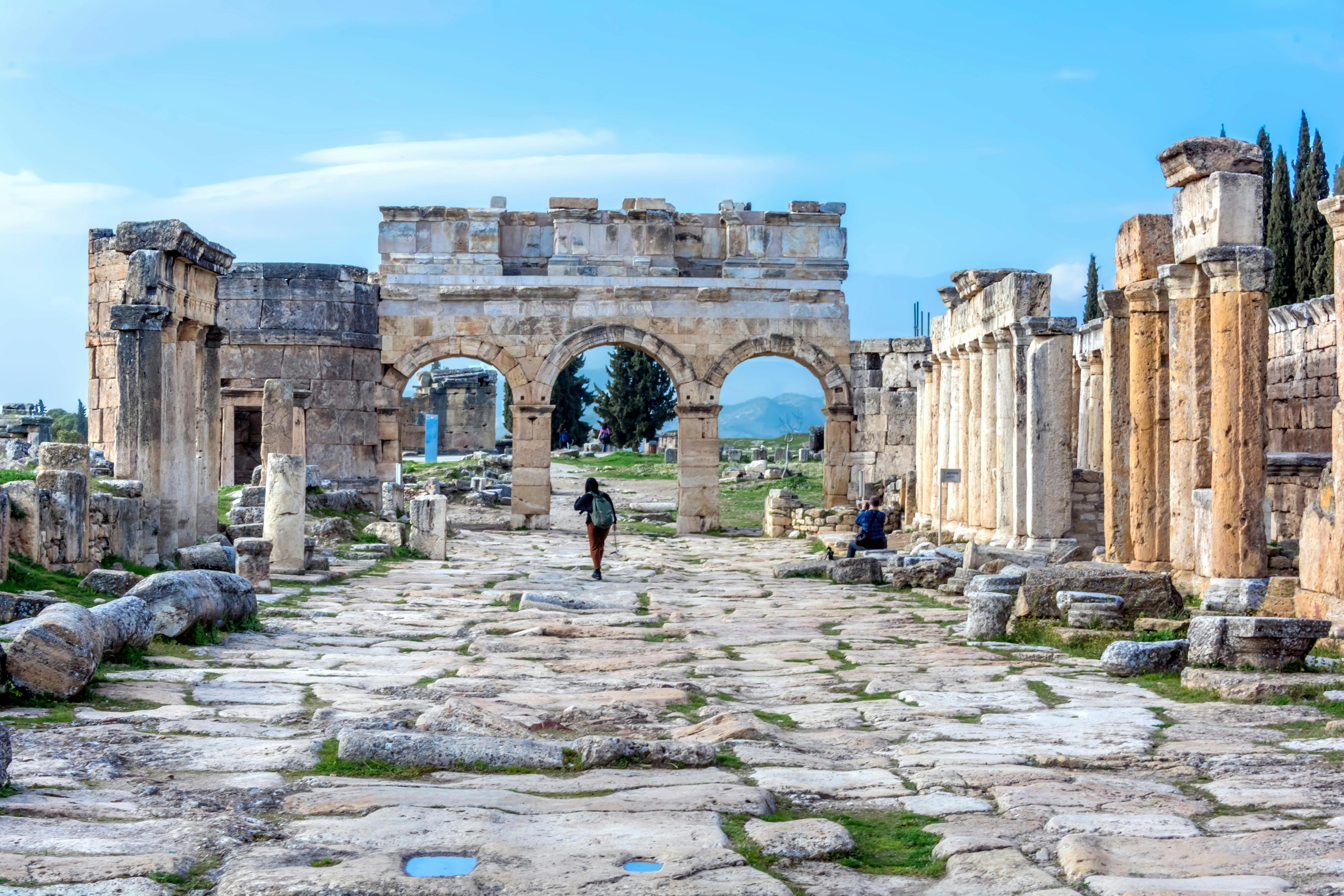 Person walking through ancient ruins with stone arches, columns, and a cobblestone path under a blue sky.