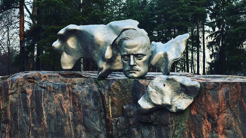 Metallic sculpture of a man's face with abstract shapes, placed on a stone platform with trees in the background.
