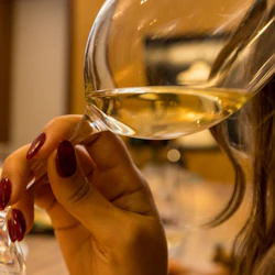 A person holding a glass of white wine with long, manicured nails in a warm, indoor setting.