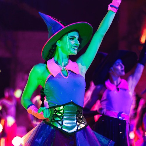 A woman dressed as a witch in a colorful costume raises her arm while smiling, with another similarly dressed woman in the background.