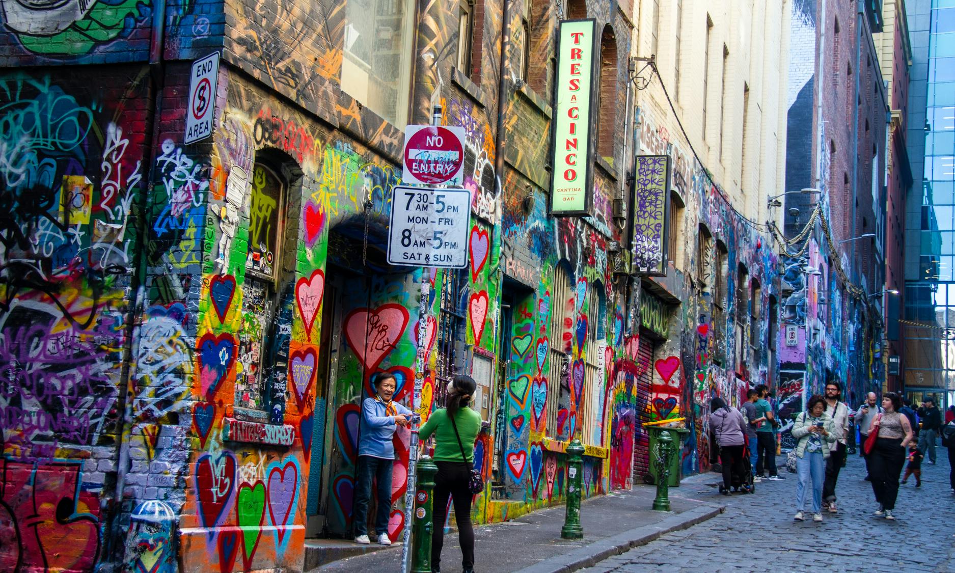 Dwie osoby stojące w pokrytej graffiti alejce o żywych sercach. Widoczne są znaki uliczne oraz znak „Tres Acinco”.