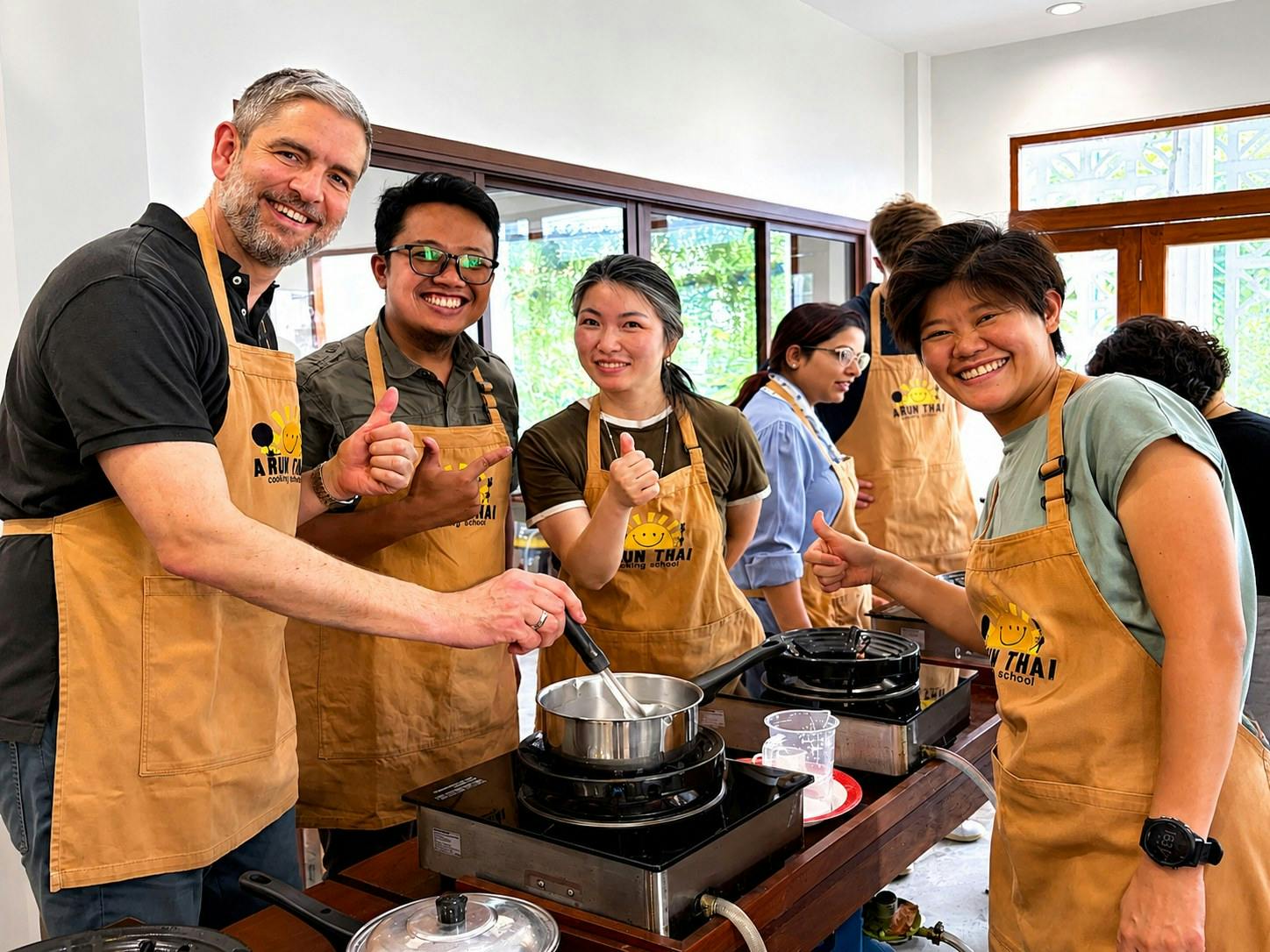 A group of people wearing aprons smile and give thumbs up while cooking at individual stovetops in a bright kitchen.