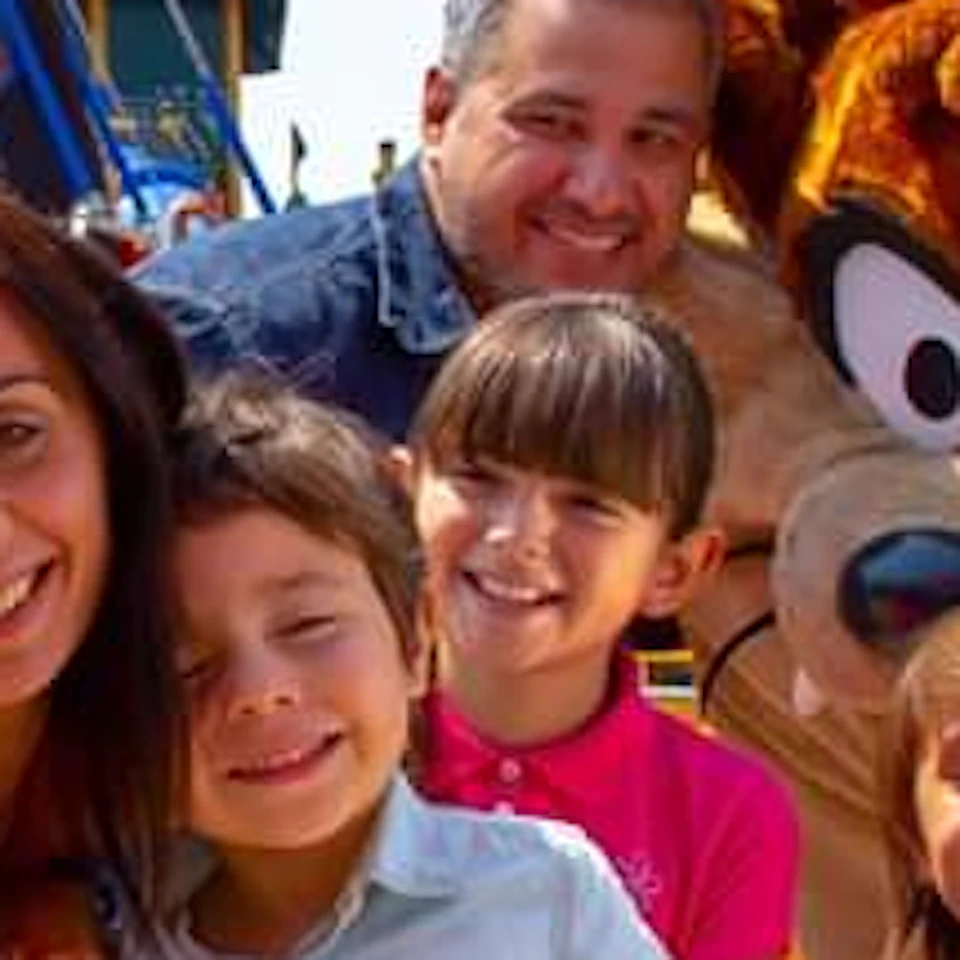 A family takes a selfie with a costumed character at an amusement park, with colorful rides and attractions visible in the background.