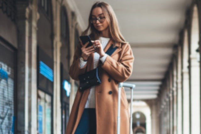 Uma pessoa de casaco castanho e óculos usa um smartphone enquanto puxa uma mala num passeio coberto.