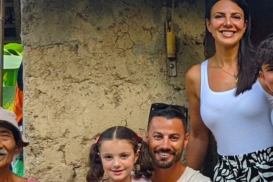A family of four posing with an elder man outside a rustic house surrounded by lush greenery.