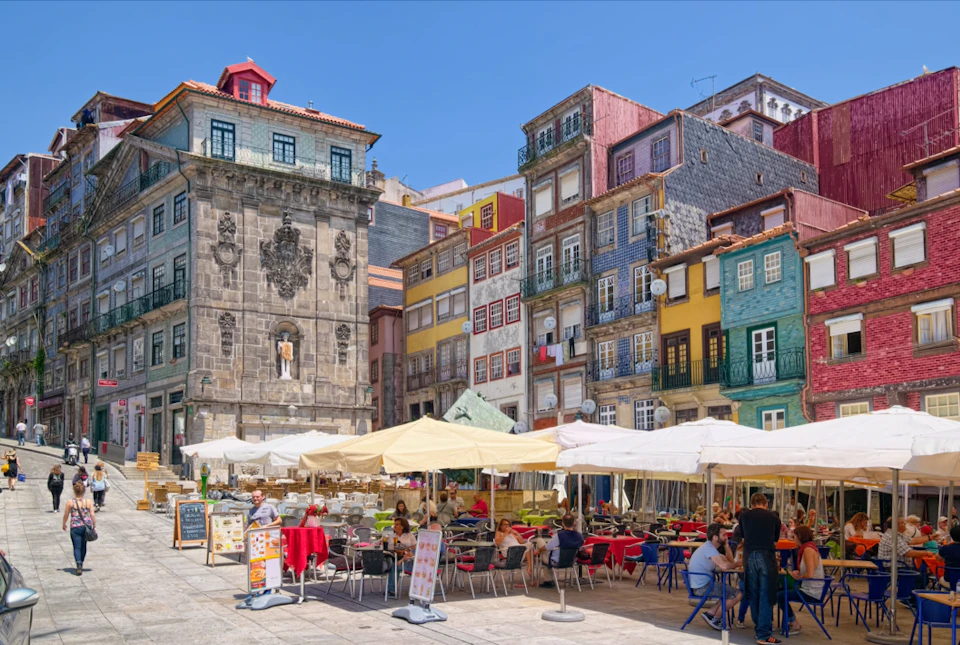 Outdoor café with people dining under umbrellas, surrounded by colorful, multi-story historic buildings on a sunny day.