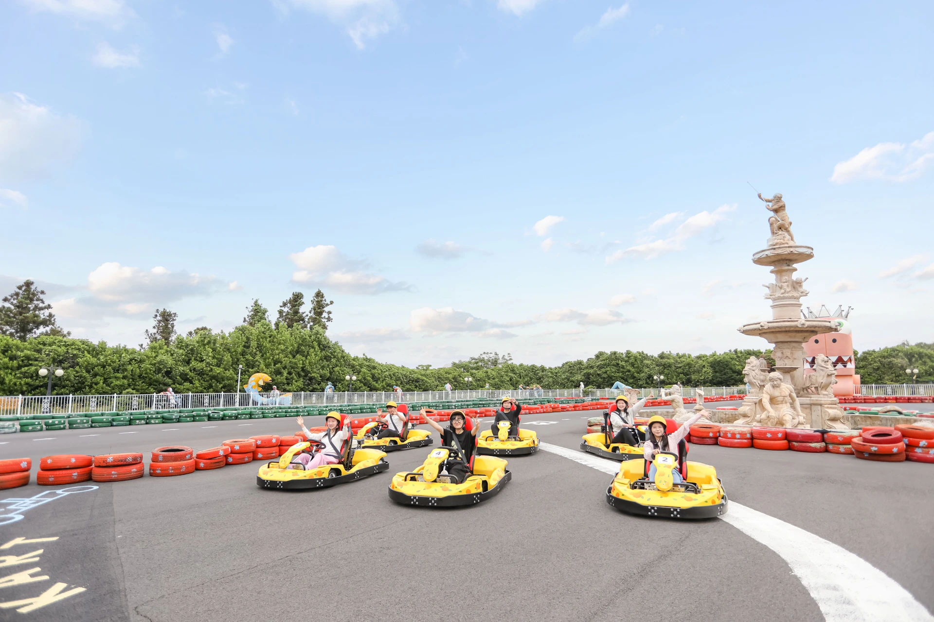 People driving yellow go-karts on a track under a clear sky, surrounded by trees and a decorative fountain.