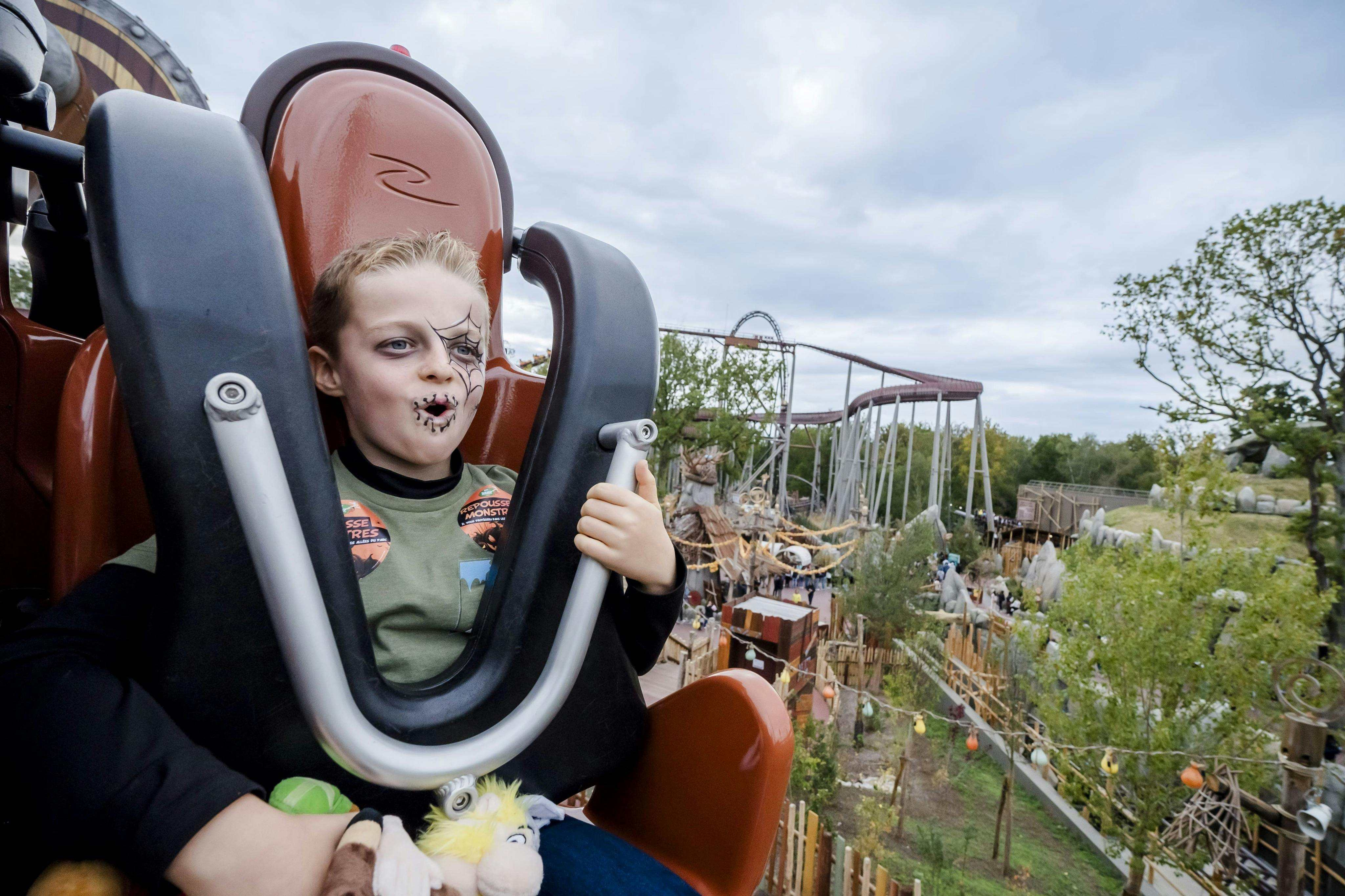 フェイスペイントとおもちゃを持った子供がジェットコースターの座席に座り、遊園地の乗り物が背景に表示されています。