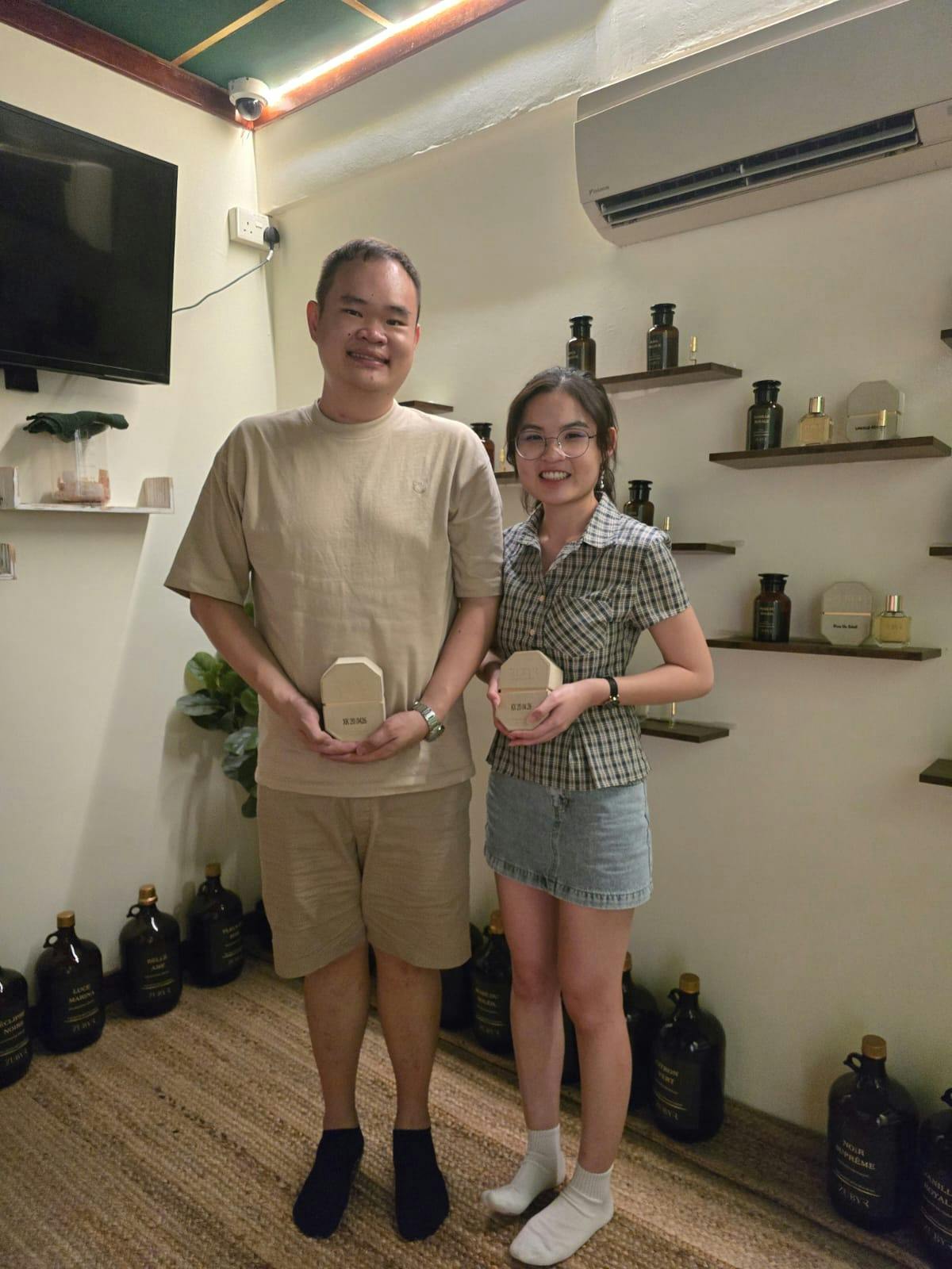 Two people stand smiling indoors, each holding a small item. Shelves with bottles and a TV are visible in the background.