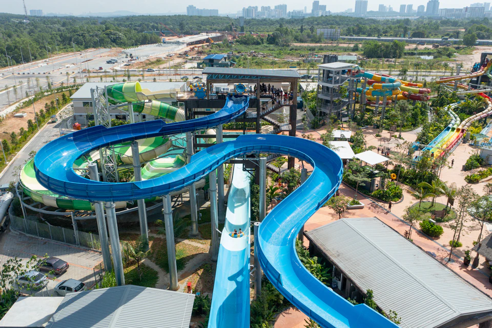 A large, winding blue water slide at a water park with people on it, surrounded by other slides and greenery. Cityscape in the distance.