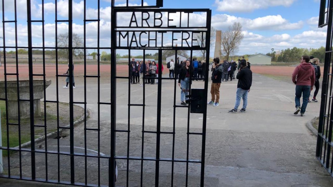 En gruppe mennesker står bag en jernport med inskriptionen "ARBEIT MACHT FREI." Himlen er delvist overskyet.