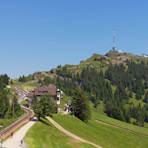 En vacker utsikt över en sluttande järnväg som leder till en bergstopp med hus, tallar och ett högt kommunikationstorn under en klarblå himmel.