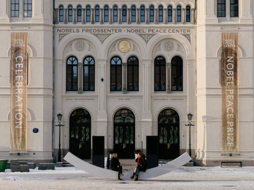 两个人坐在诺贝尔和平中心外的现代长凳上，横幅上写着“庆祝”和“诺贝尔和平奖”。