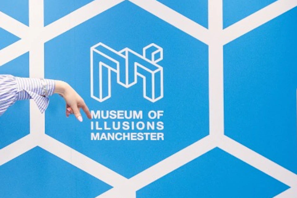 A person stands in a blue room with geometric wall patterns and a sign reading 'Museum of Illusions Manchester.'