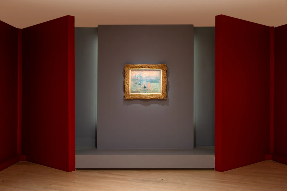 A framed painting displayed on a gray wall, flanked by two red panels in a gallery setting with wooden flooring.