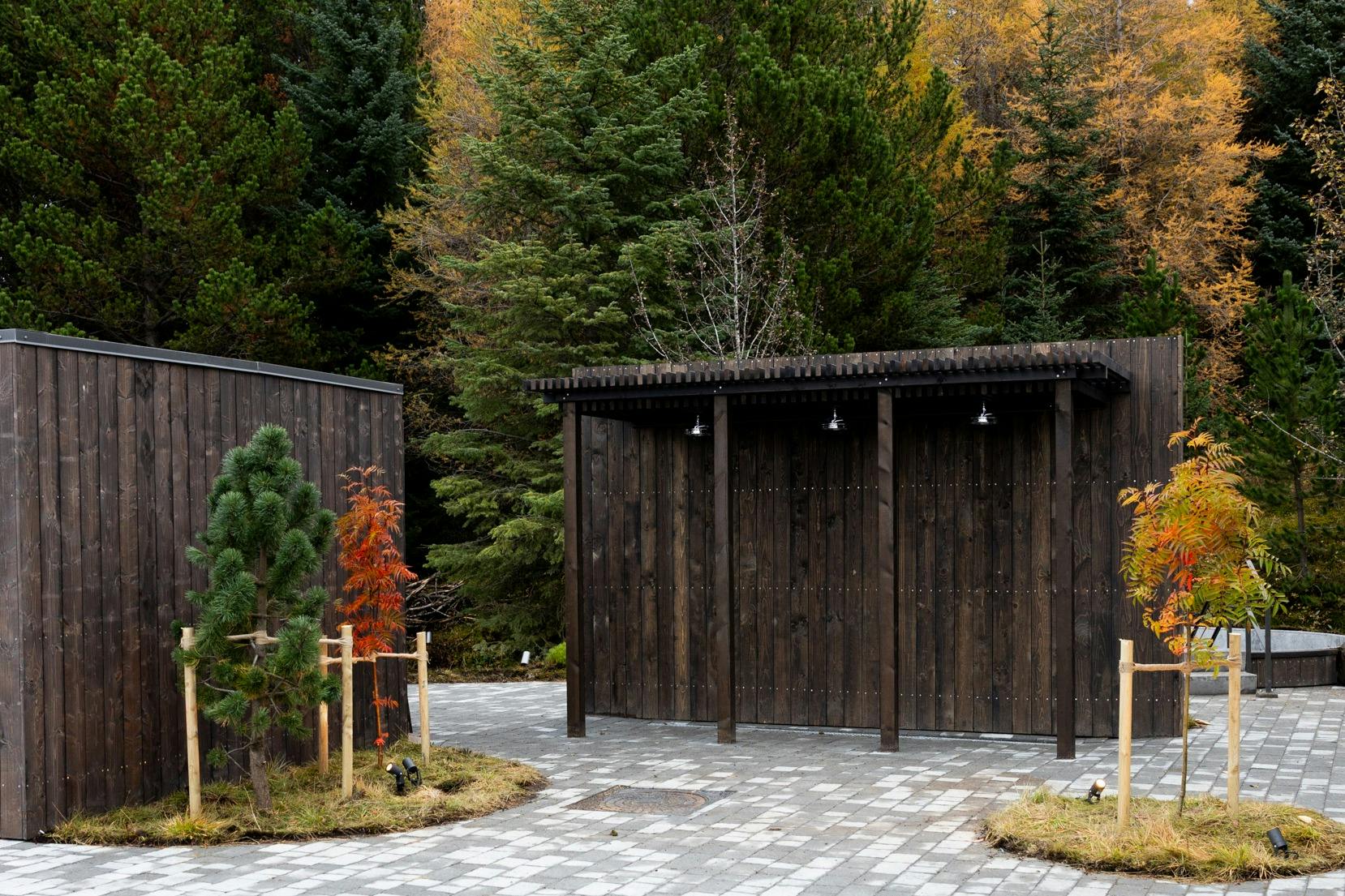 A dark wooden shelter with three hanging lights stands beside a pathway surrounded by lush green and autumnal trees.