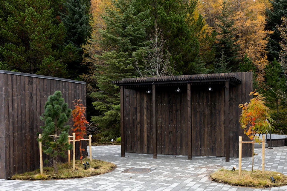 A dark wooden shelter with three hanging lights stands beside a pathway surrounded by lush green and autumnal trees.