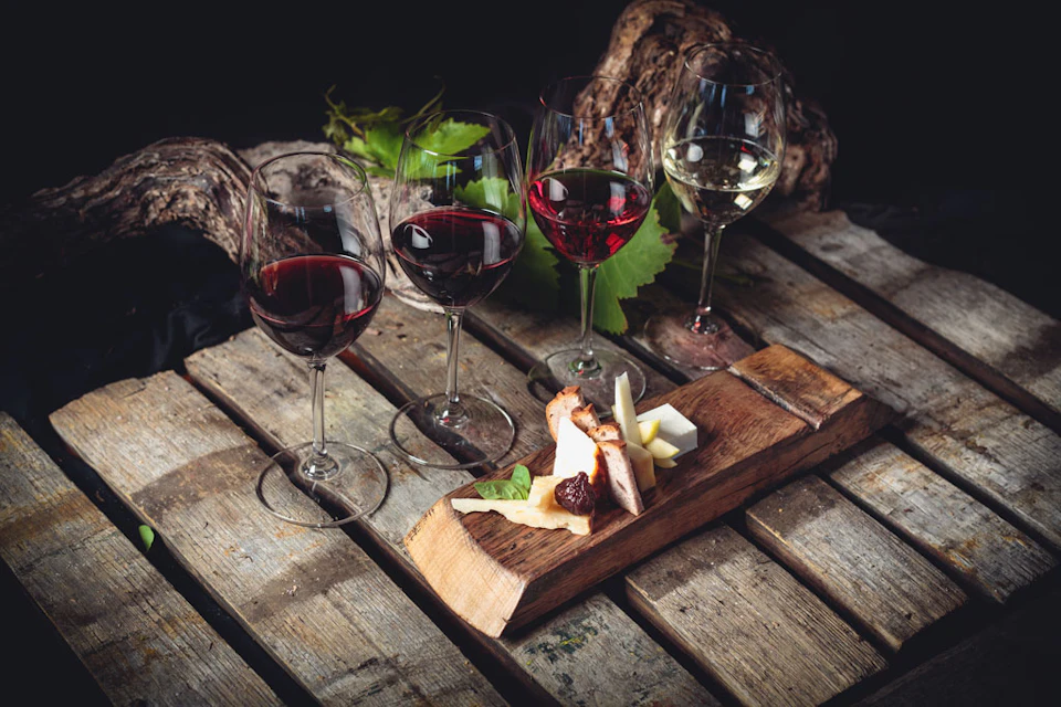 Four wine glasses with different wines on a wooden table, accompanied by a cheese and meat platter with leafy greens.