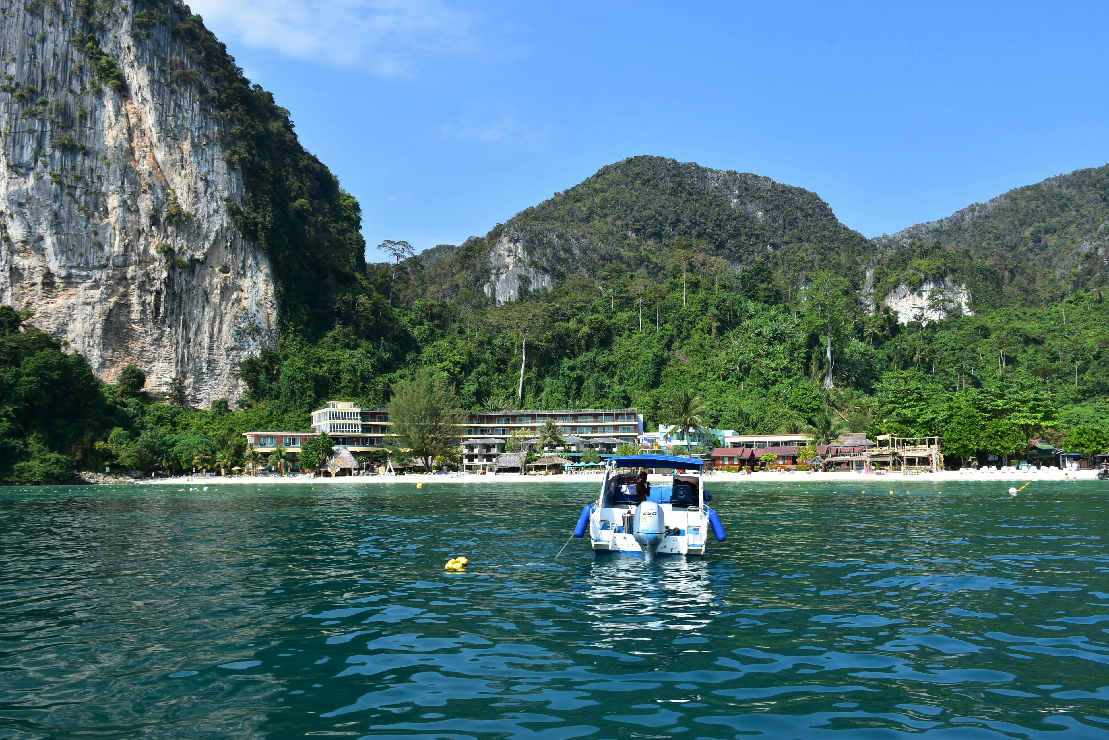 Μια βάρκα σε ήρεμα πράσινα νερά με φόντο καταπράσινα βουνά και κτίρια στην ακτή κάτω από έναν καταγάλανο ουρανό.