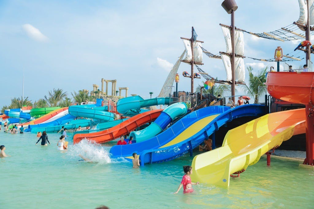 Children playing in a water park with colorful slides and a pirate ship. Some kids are sliding down while others are swimming.
