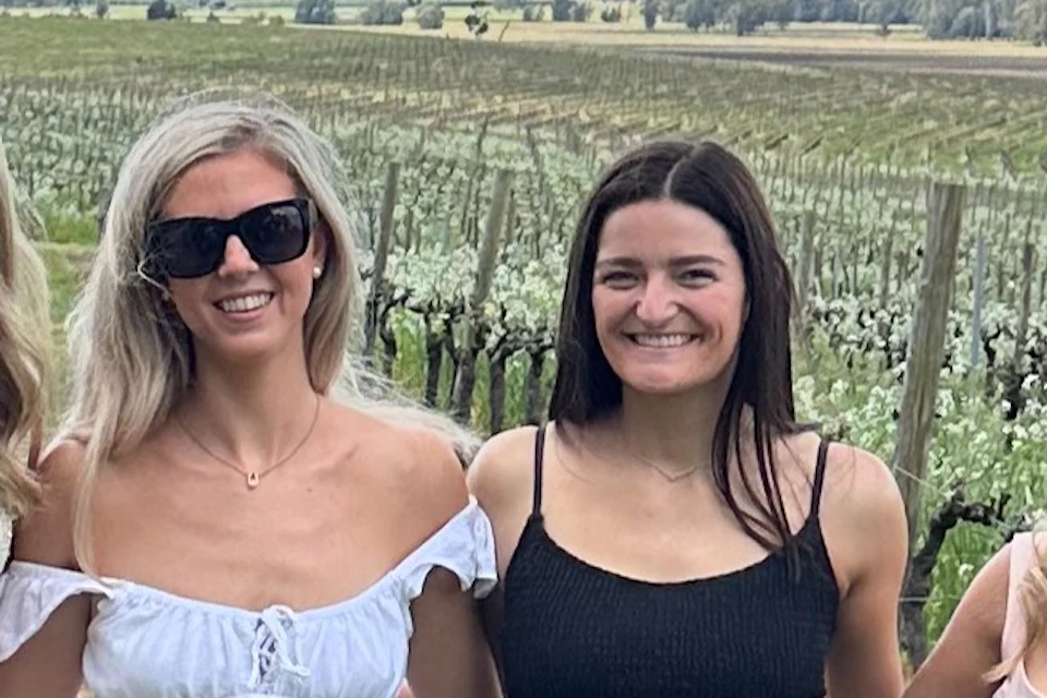 A group of six women posing outdoors in a vineyard on a sunny day, dressed in casual summer dresses, with a scenic background.