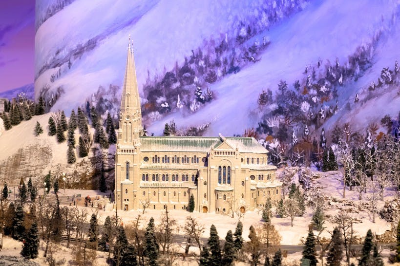 En detaljeret model af en stor kirke med et højt spir, omgivet af sne og træer, sat mod en malet bjergrig baggrund.