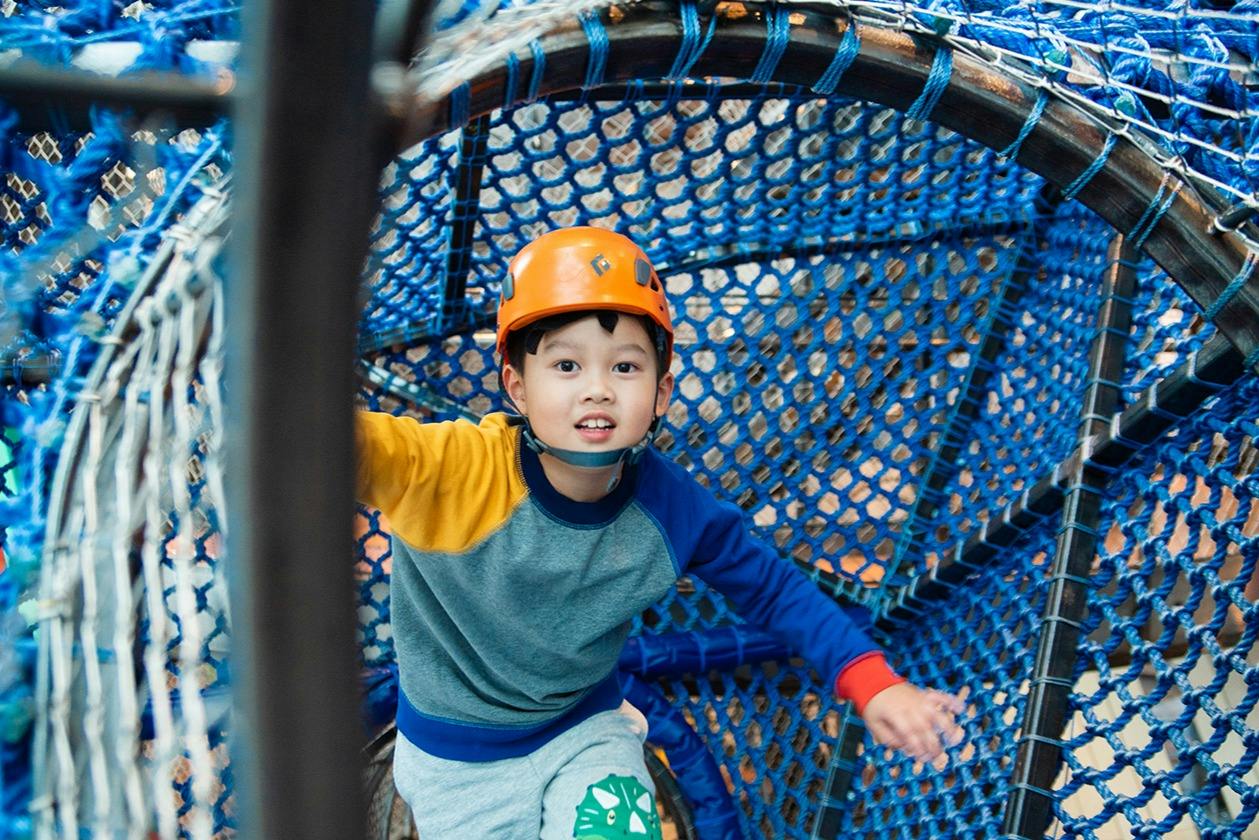 A child wearing an orange helmet climbs inside a blue net structure in an indoor play area.