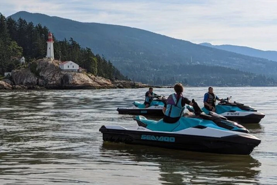 Three people on jet skis floating near a rocky shore with a white lighthouse and house, surrounded by trees, under a clear sky.
