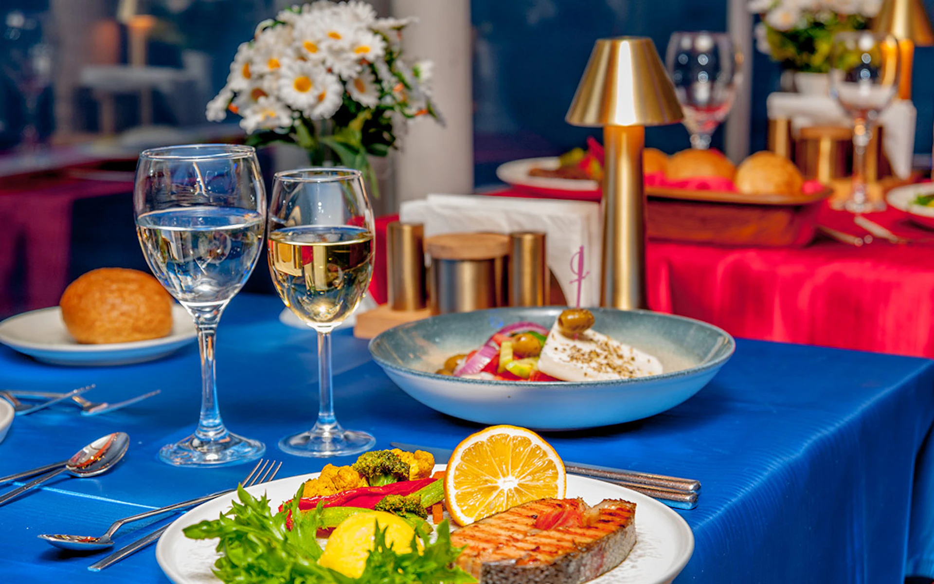 A table set with wine glasses, grilled salmon, vegetables, and salad. Flowers and a small lamp are in the background.