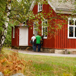 Två personer står nära ingången till ett rött trähus omgivet av grönt och gult lövverk.