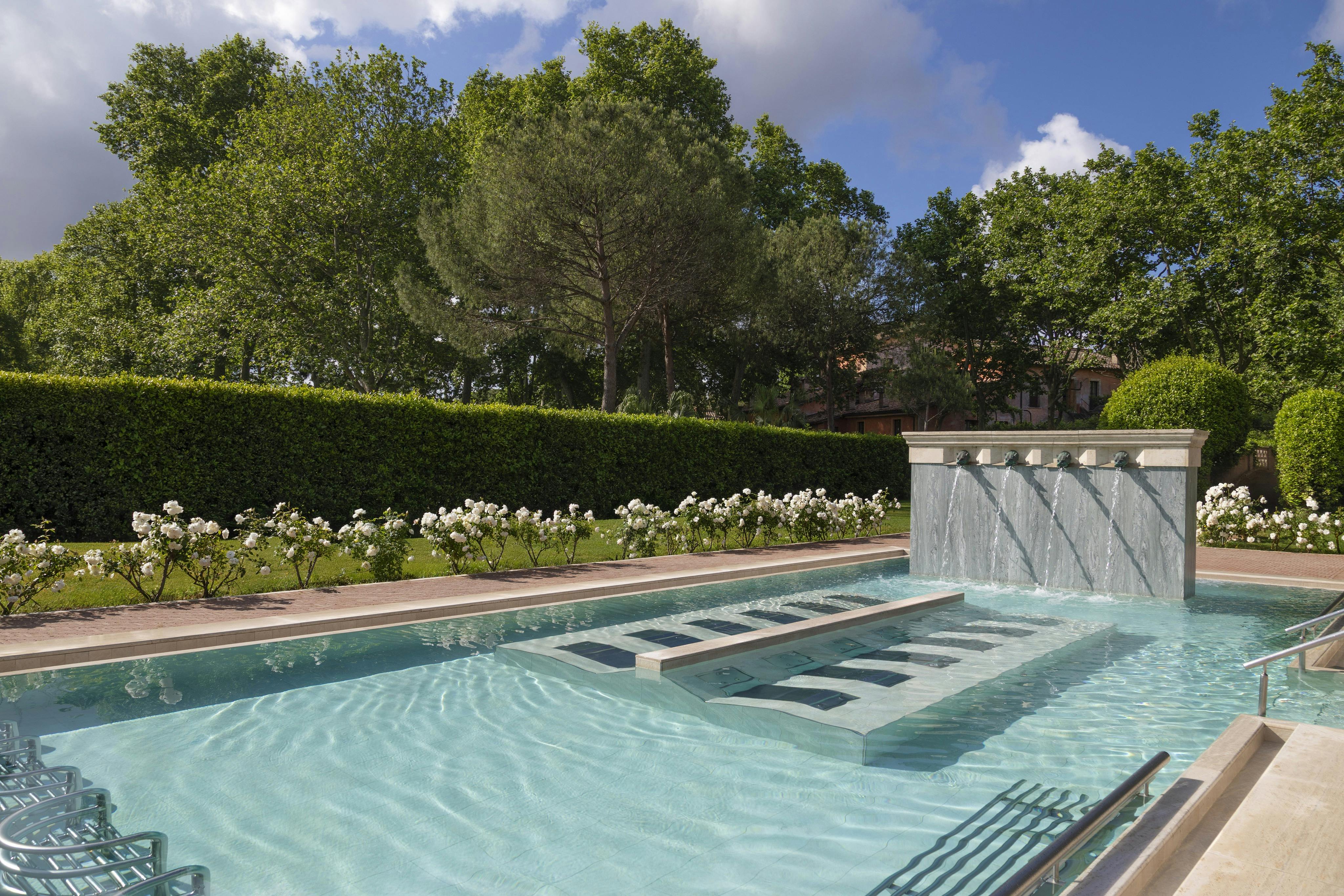 A serene outdoor pool with a waterfall feature, surrounded by greenery and flowers under a partly cloudy sky.