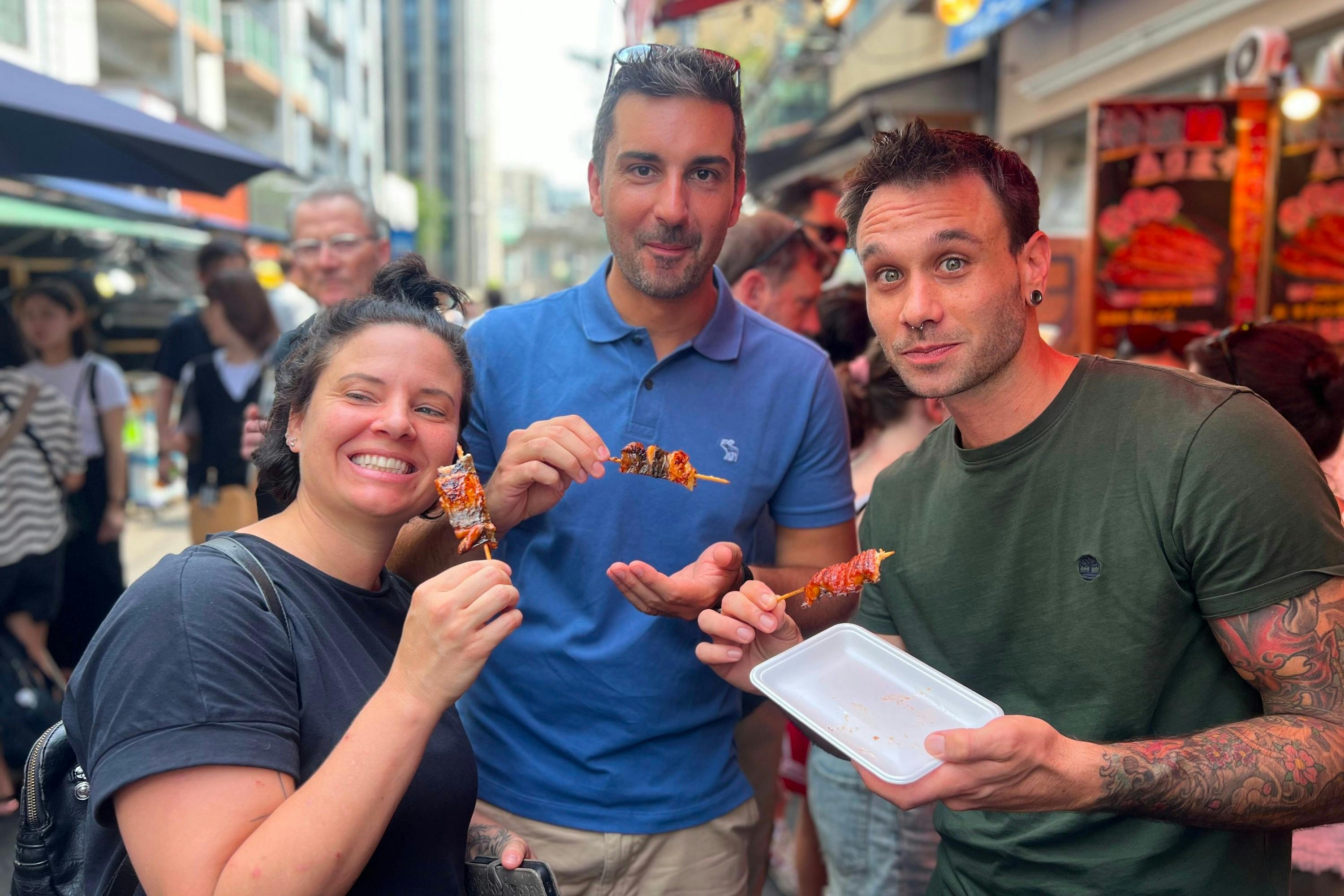 Three people standing outdoors, smiling and holding meat skewers, with a city street and crowd in the background.