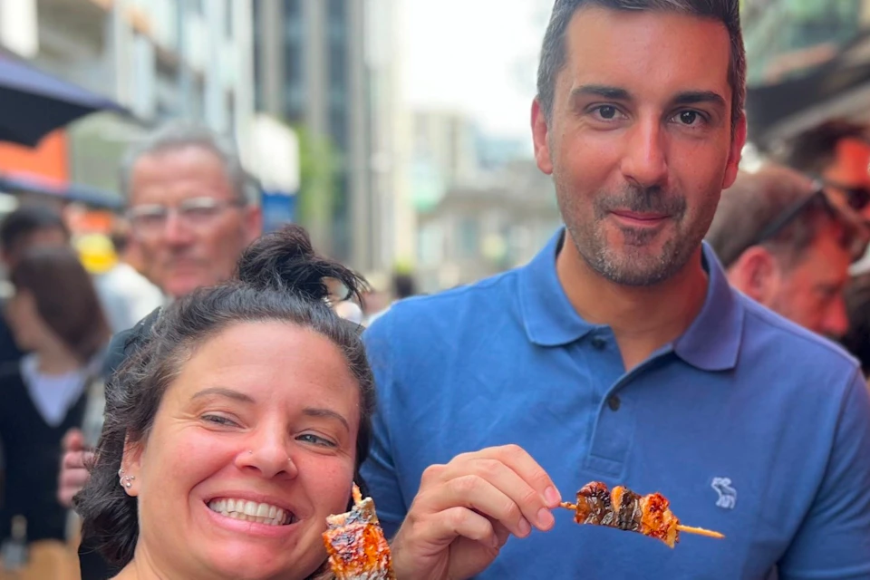 Three people standing outdoors, smiling and holding meat skewers, with a city street and crowd in the background.