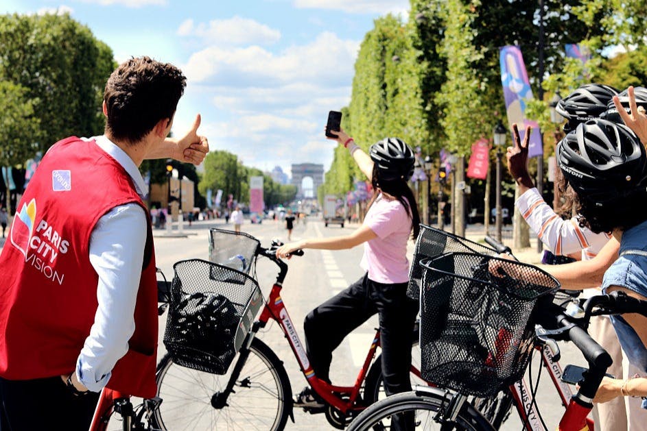 Three cyclists on a tree-lined street take a photo with the Arc de Triomphe in view. One person gives a thumbs-up.
