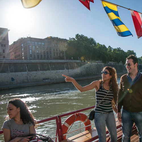 Människor på en båt med färgglada flaggor ovanför, som njuter av en solig dag på en flod nära en stad. En kvinna pekar mot stranden.
