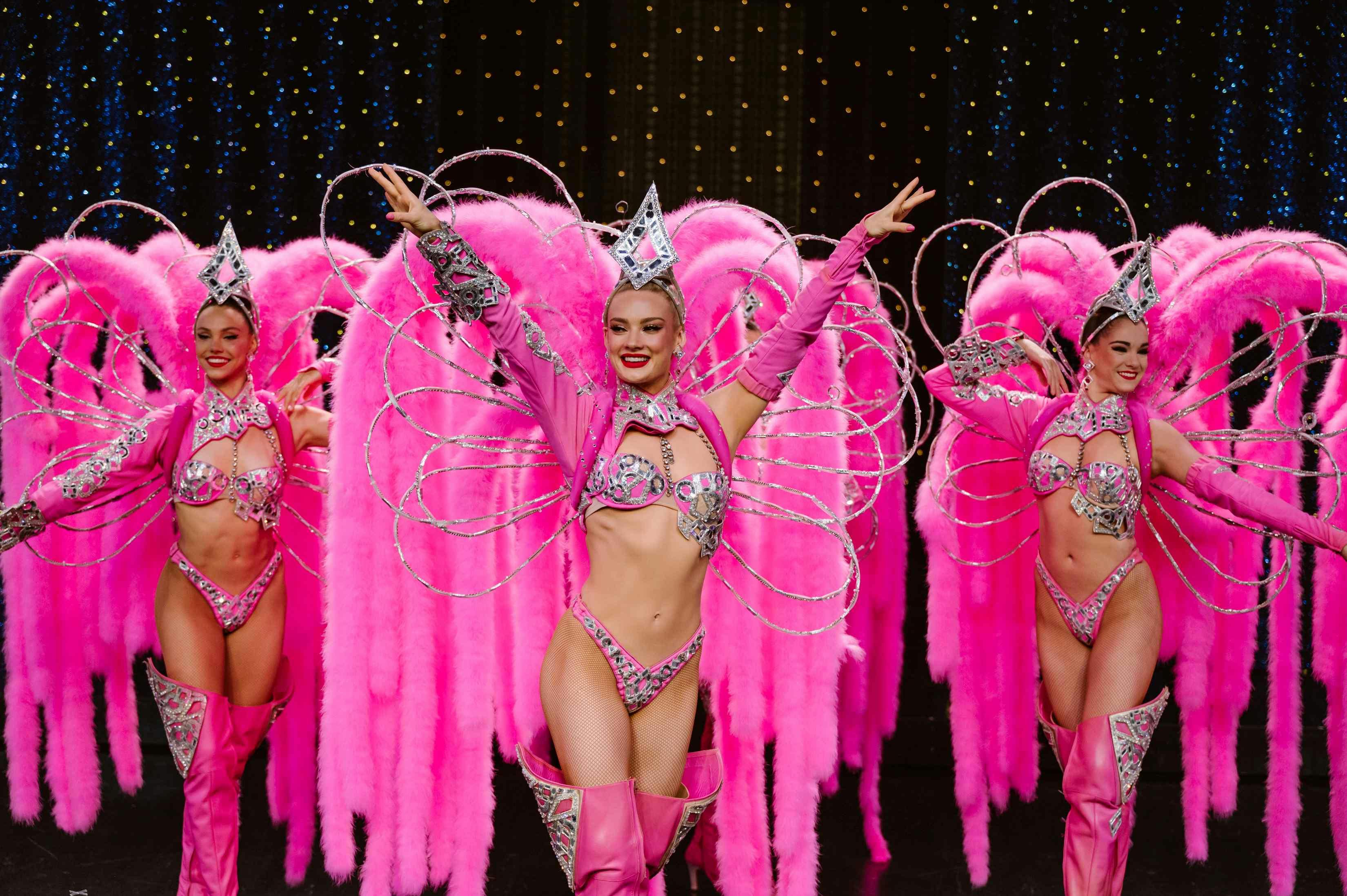 Performers in vibrant pink costumes with feathers and sparkling decorations dance on a stage with a dark background.