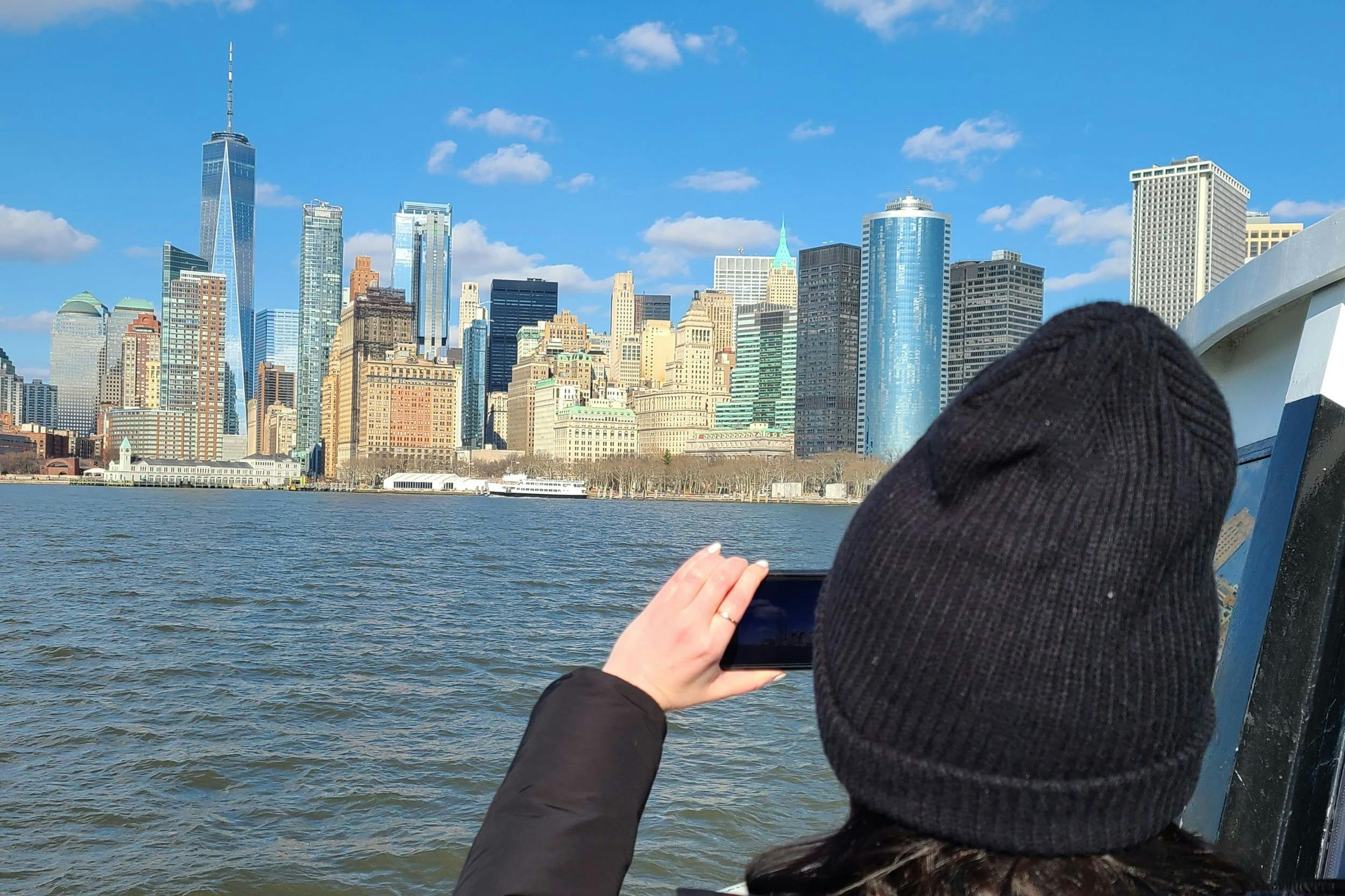 Eine Person mit schwarzem Hut fotografiert an einem klaren Tag mit blauem Himmel die Skyline einer Stadt über dem Wasser.