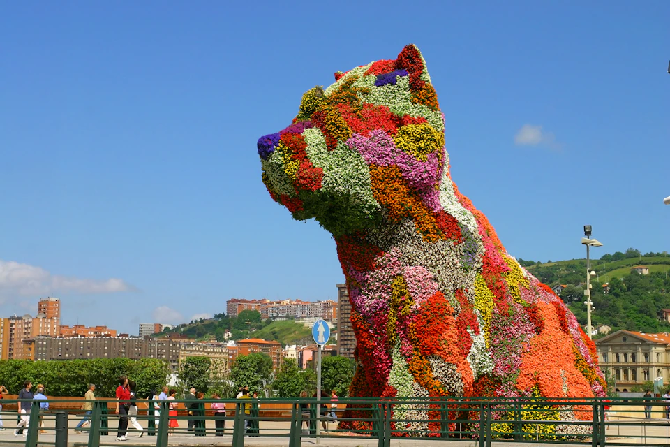 Large floral sculpture of a dog with various colorful flowers set in an urban park with people walking and buildings in the background.